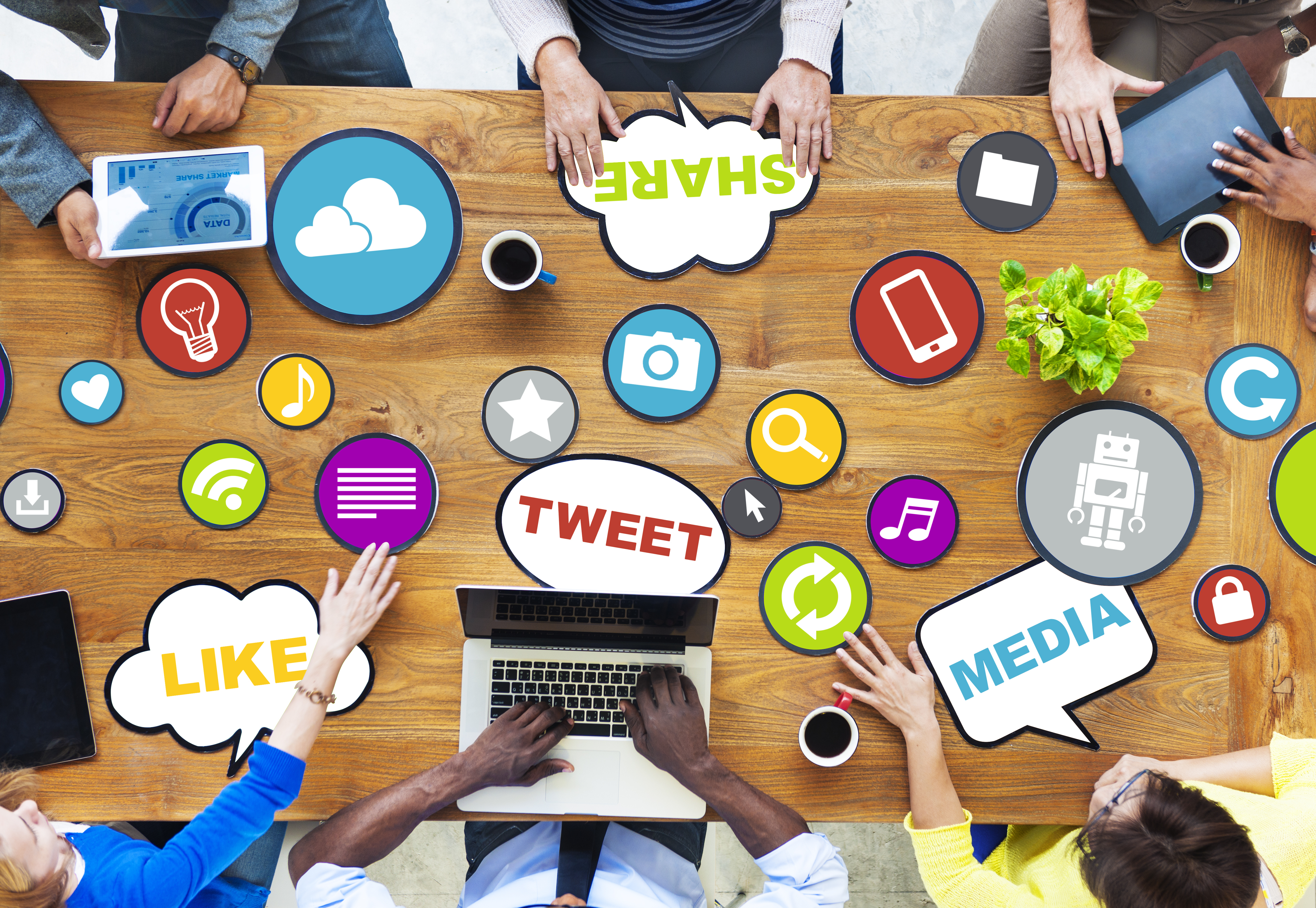 Six people sitting around a wooden table working on various devices surrounded by different place-mats with different icons and the words: share, like, tweet, and media. 